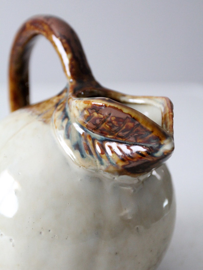 May include: A white ceramic pitcher with a brown handle and a brown leaf design. The pitcher has a glossy finish and a slightly textured surface.