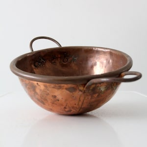May include: A small, copper bowl with two wire handles. The bowl has a hammered finish and shows signs of age.