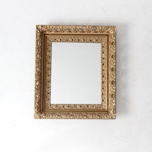 May include: Square gold-colored mirror with an ornate, detailed frame. The frame has a textured, vintage appearance with intricate patterns and designs. The mirror's reflective surface is clear and smooth, contrasting with the textured frame.