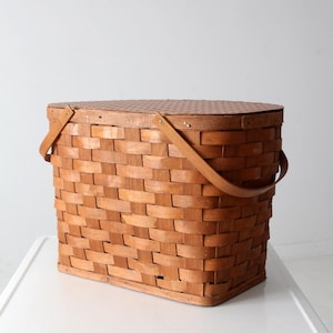 以下が含まれることがあります： 木製のハンドルと蓋が付いた長方形のピクニックバスケット。バスケットは、織り込まれた茶色の木のストリップで作られています。蓋は明るい茶色で、テクスチャパターンがあります。ハンドルは、明るい茶色の革のような素材で作られています。