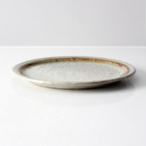 May include: A small, round, white ceramic plate with a brown rim. The plate has a speckled, textured surface.