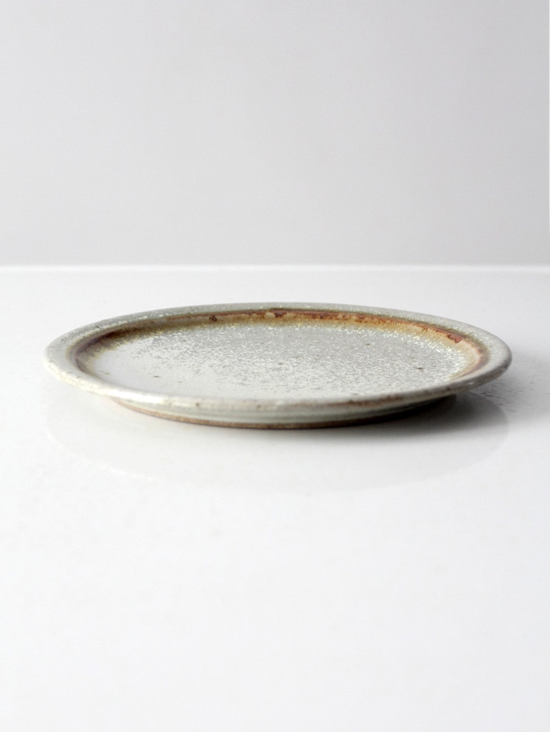May include: A small, round, white ceramic plate with a brown rim. The plate has a speckled, textured surface.