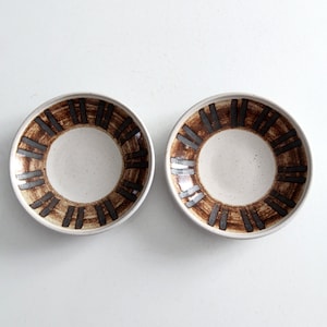 May include: Two white ceramic bowls with brown and silver geometric designs. The bowls are empty and sitting on a white surface.