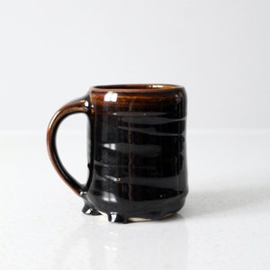 May include: A brown and black ceramic mug with a handle. The mug has a textured, slightly uneven surface.