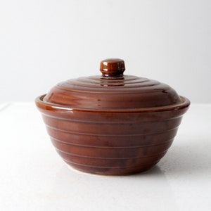 Puede incluir: Un tazón de cerámica marrón con tapa. El tazón tiene una forma redondeada y está decorado con una serie de líneas horizontales.