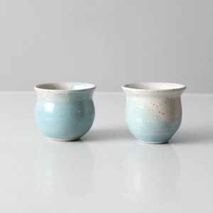 May include: Two small, light blue ceramic cups with a white rim and speckles. The cups are round and have a slight curve to them.