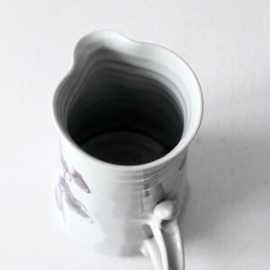 May include: White ceramic pitcher with a blue and gray floral design. The pitcher has a unique, slightly uneven shape and a handle.