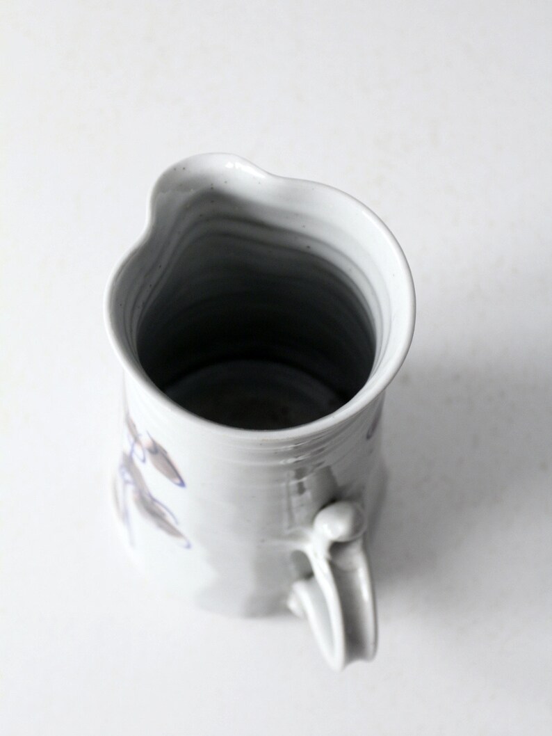 May include: White ceramic pitcher with a blue and gray floral design. The pitcher has a unique, slightly uneven shape and a handle.