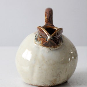 May include: A small, round ceramic pitcher with a brown glaze and a white glaze. The pitcher has a handle and a spout.