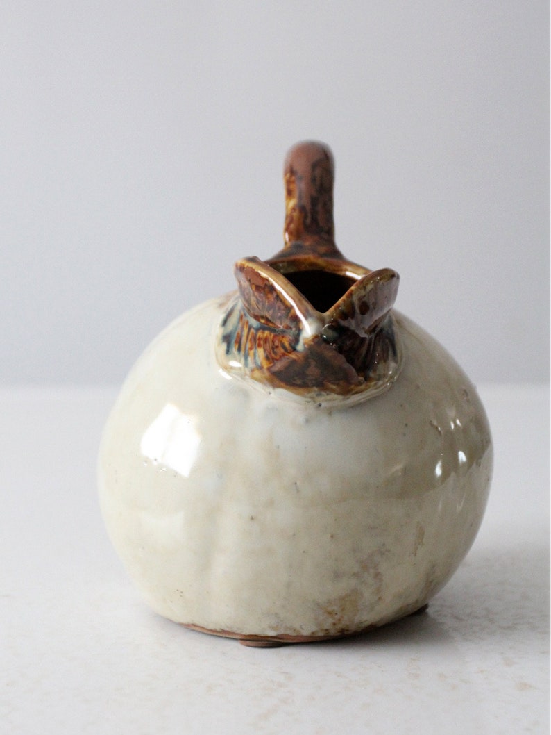 May include: A small, round ceramic pitcher with a brown glaze and a white glaze. The pitcher has a handle and a spout.