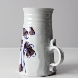May include: White ceramic vase with a brown and blue floral design and a handle. The vase has a textured surface and a slightly flared opening.