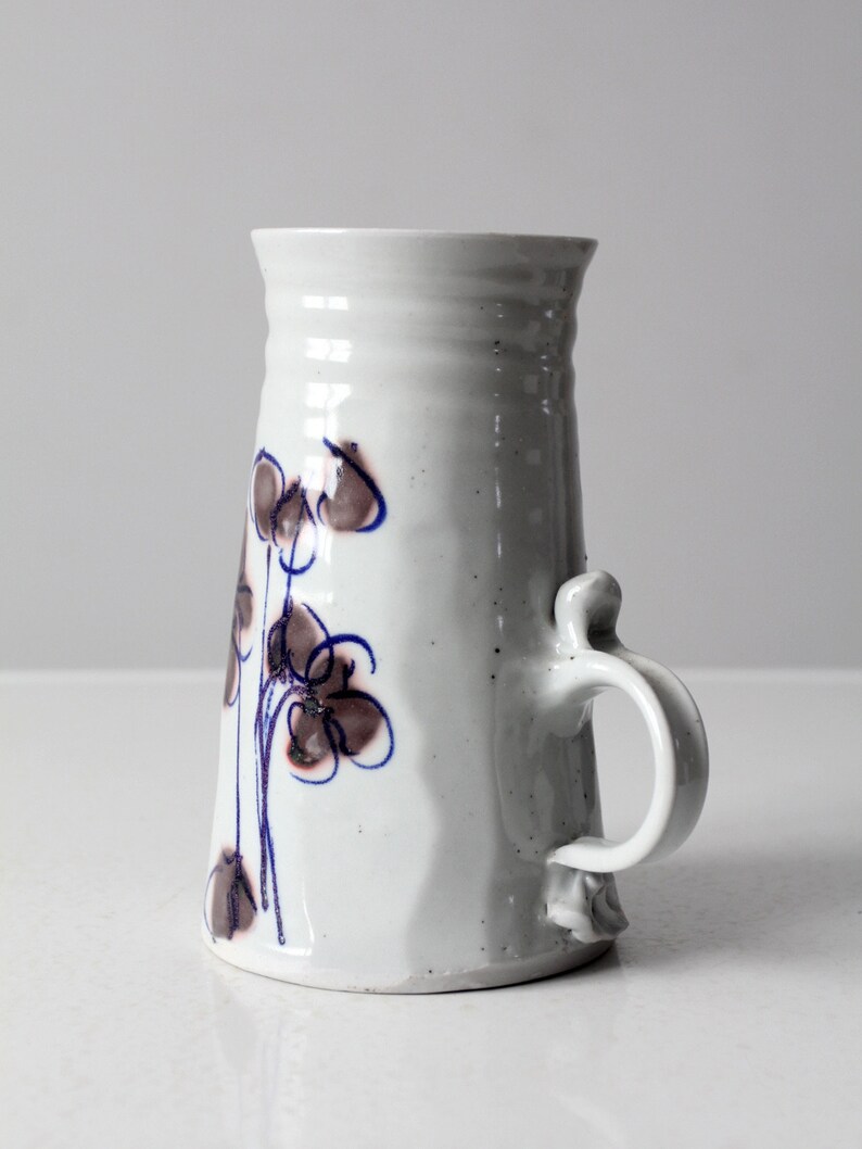 May include: White ceramic vase with a brown and blue floral design and a handle. The vase has a textured surface and a slightly flared opening.