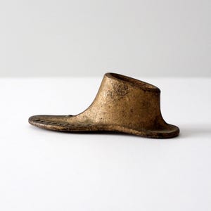 Puede incluir: Una horma de zapato antigua, hecha de metal con un acabado dorado envejecido. La forma del zapato presenta un talón acampanado y una suela plana y alargada, mostrando su diseño histórico y artesanía.