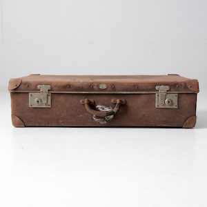 May include: A vintage brown suitcase with metal latches and a handle. The rectangular suitcase has reinforced corners and a slightly worn appearance, suggesting age and use. The metal hardware is tarnished, adding to the antique aesthetic.
