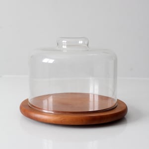 Op de afbeelding: Een heldere glazen stolp met een afgeronde bovenkant staat op een ronde, houten basis. De basis heeft een warme, bruine kleur. De stolp is ontworpen om items te bedekken en te beschermen.