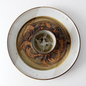 May include: A ceramic serving platter with a central dip bowl. The platter features a white rim and a brown and tan center with an abstract design. The dip bowl is tan with a white cross design.