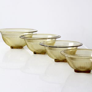 Op de afbeelding: Een set van vier amberkleurige glazen mengkommen van verschillende formaten. De kommen hebben een geribbeld ontwerp en staan op kleine, vierkante voetjes. De kommen zijn diagonaal gerangschikt, met de grootste kom achteraan en de kleinste vooraan.