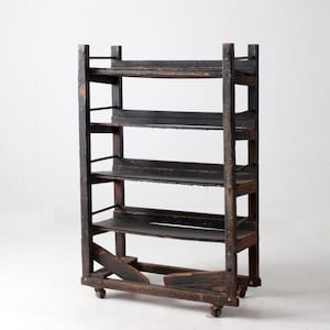 May include: A vintage, dark-stained wooden shelf unit with four shelves. The shelves are supported by a frame with vertical posts and horizontal supports. The unit has small wheels on the bottom for mobility. The wood shows signs of wear, giving it a rustic appearance.