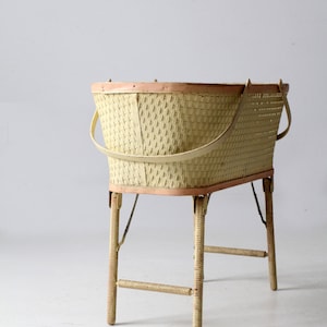 May include: A vintage wicker baby bassinet with a light pink trim and white painted legs. The bassinet has two handles and a woven design.