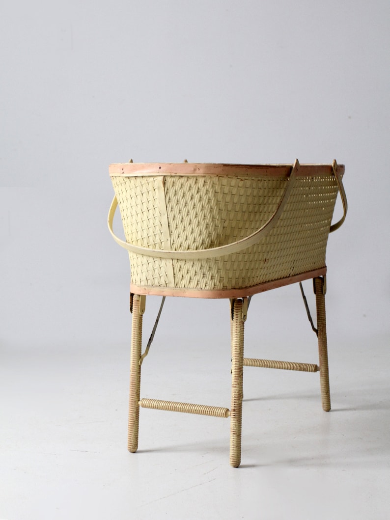 May include: A vintage wicker baby bassinet with a light pink trim and white painted legs. The bassinet has two handles and a woven design.