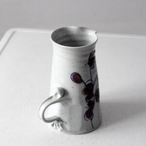 May include: White ceramic pitcher with a blue and brown floral design and a unique handle.
