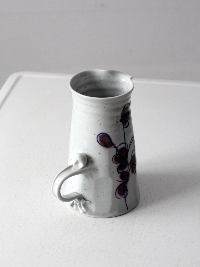 May include: White ceramic pitcher with a blue and brown floral design and a unique handle.
