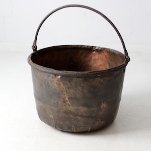 May include: A rustic, antique metal cauldron with a dark, aged patina. The cauldron has a rounded body and a sturdy, arched handle. The metal shows signs of wear and discoloration, adding to its vintage charm. The interior appears dark and possibly coated.
