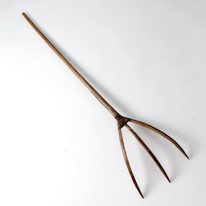 May include: A long, wooden pitchfork with three tines. The handle is a single piece of wood, and the tines are curved and pointed. The pitchfork is a light brown color and appears to be handmade.