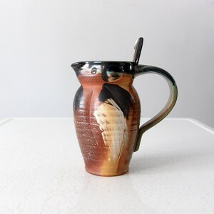 Könnte beinhalten: Ein brauner und schwarzer Keramik Krug mit einem weißen und gelben Design. Der Krug hat einen Henkel und einen kleinen Ausguss.