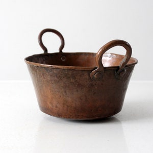 May include: A small, antique copper pot with two loop handles. The pot has a hammered finish and is slightly dented.