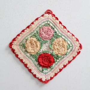 Puede incluir: Un salvamanteles cuadrado hecho a mano con ganchillo, con cuatro diseños florales en tonos rosa, amarillo y rojo, rodeados de hojas verdes. El salvamanteles tiene un borde rojo festoneado y un lazo para colgar.