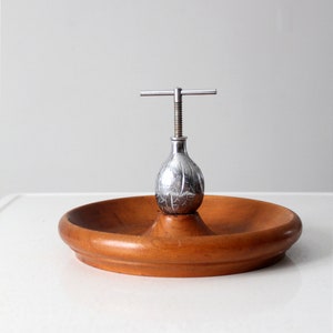 Op de afbeelding: Een zilveren notenkraker met een houten voet. De notenkraker is peer-vormig en heeft een decoratief ontwerp. De voet is rond en heeft een gladde afwerking.