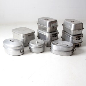 May include: A set of vintage aluminum cookware with lids and handles. The cookware is stacked and arranged on a white surface.