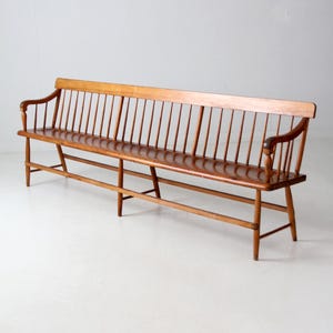 May include: A long, wooden bench with a slatted back and armrests. The bench is made of light brown wood and has a glossy finish. The back of the bench features vertical spindles. The bench is supported by four legs and two horizontal supports.