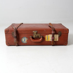 May include: A vintage brown leather suitcase with brass hardware and leather straps. The suitcase has a sticker that says "Tourists" and another sticker that says "Amerika Linie" with a letter "R" inside a circle.