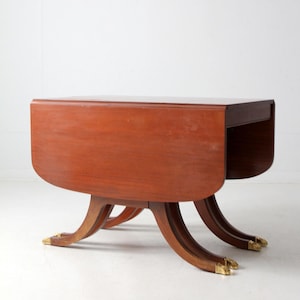 May include: A vintage mahogany drop-leaf table. The table has a rectangular top with rounded edges and a drop-leaf design. The table is supported by four curved legs with brass claw feet. The table is approximately 30 inches long when closed.