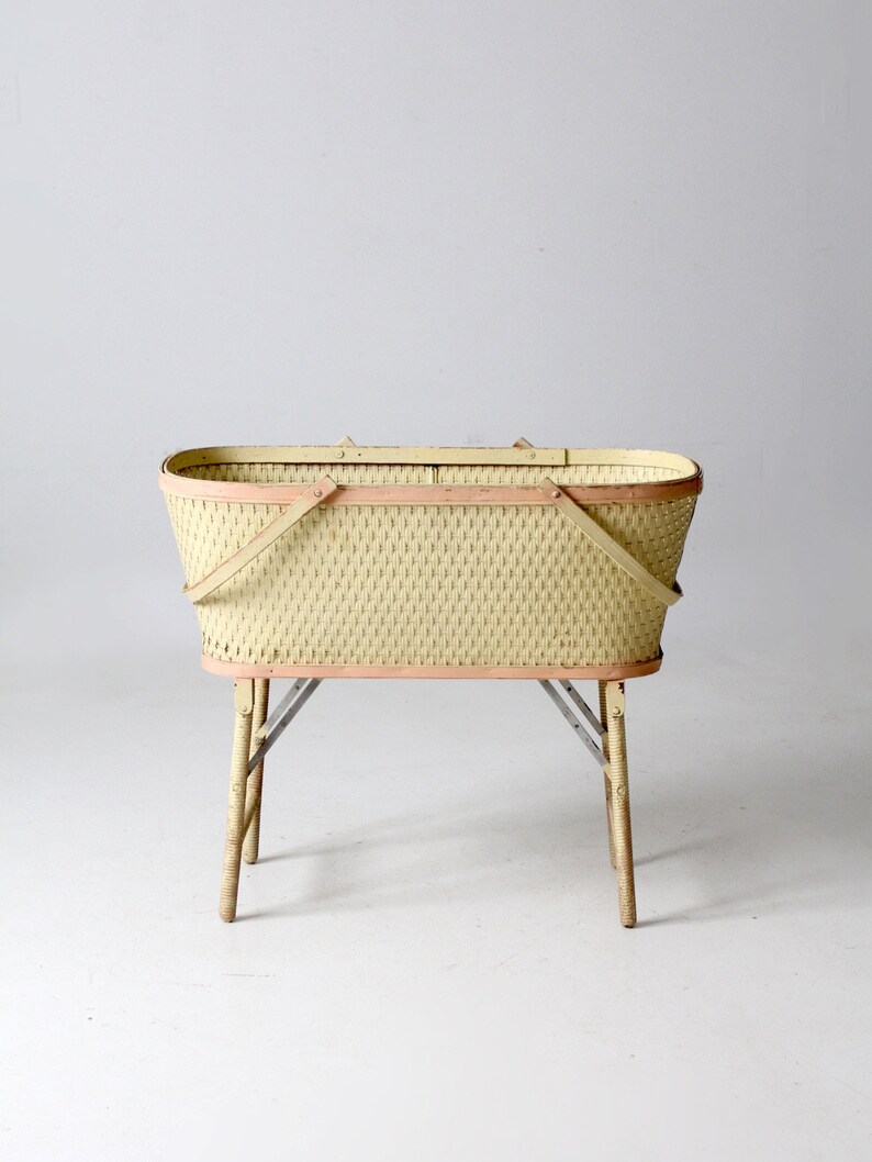 May include: A vintage wicker basket with a light yellow finish and pink trim. The basket has a folding handle and sits on four wooden legs.