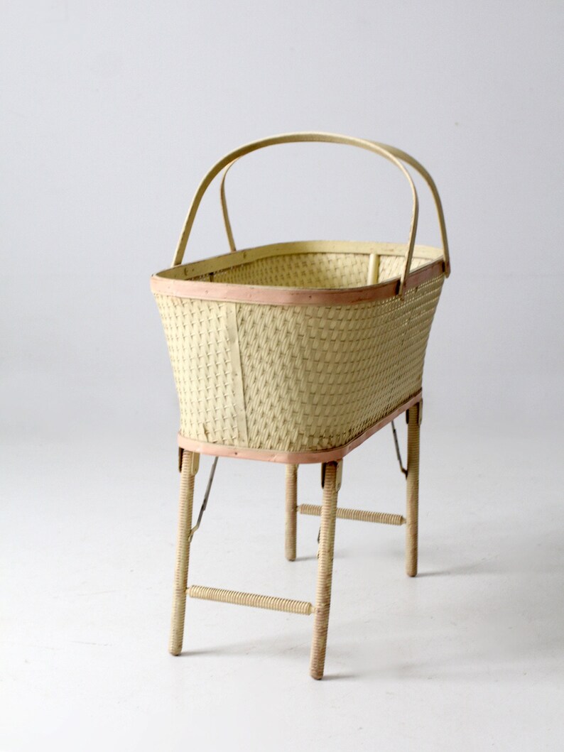 May include: A white wicker baby bassinet with a pink trim and folding legs. The bassinet has a handle on the top.