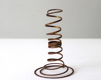 May include: A rusty metal spring coil with a circular base. The spring is coiled vertically and the base is made of a single, continuous coil of wire.