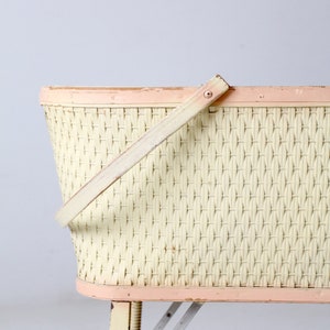 May include: A vintage wicker baby bassinet with a pink painted rim and a wooden handle. The bassinet has a white wicker weave and sits on a set of folding metal legs.