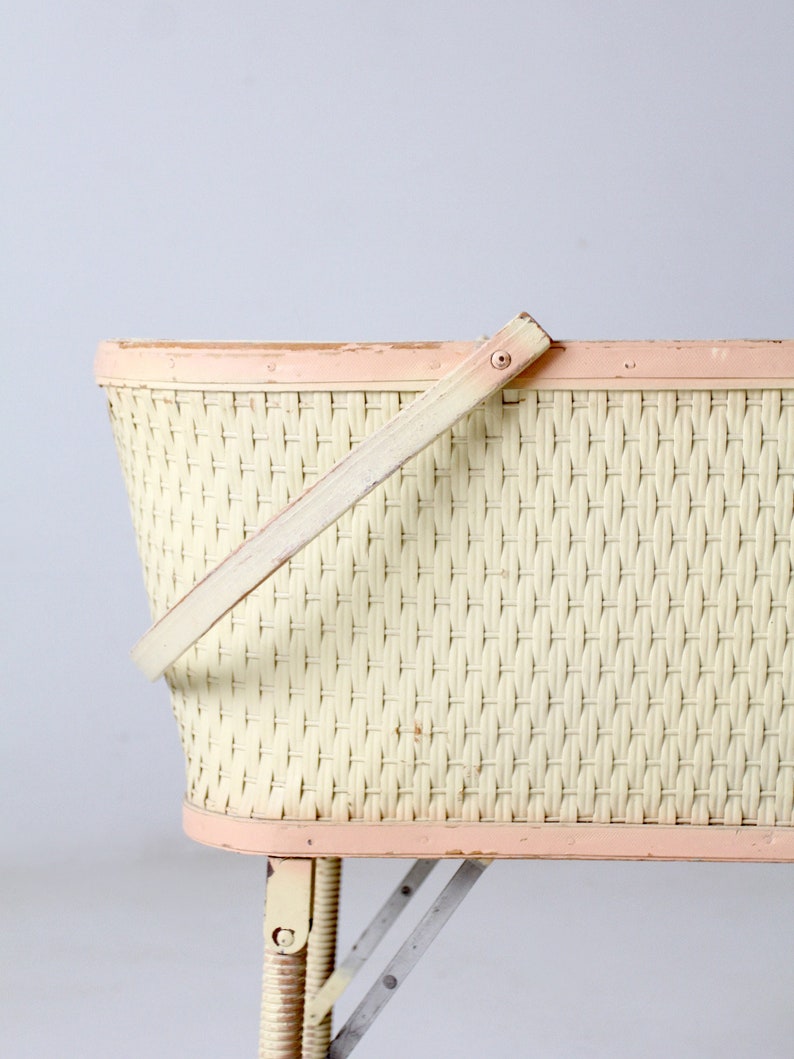 May include: A vintage wicker baby bassinet with a pink painted rim and a wooden handle. The bassinet has a white wicker weave and sits on a set of folding metal legs.