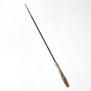 May include: Vintage fishing rod with a dark rod and a cork handle. The rod has metal guides and a metal reel seat. The rod is angled diagonally against a white background.