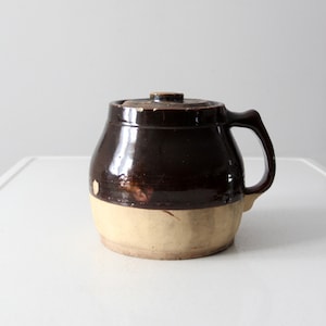May include: A brown and cream glazed stoneware crock with a lid and a handle. The crock has a rustic, vintage look.