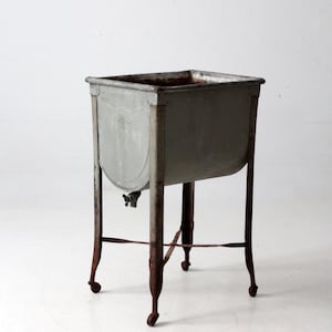 May include: An antique metal washstand with a rectangular basin and a spigot. The stand has four legs with small wheels and a crossbar for stability. The metal has a weathered, gray finish with rust.