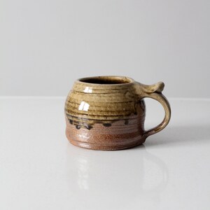 May include: A brown and tan ceramic mug with a unique handle. The mug has a brown glaze with a tan rim and a brown drip glaze around the bottom.