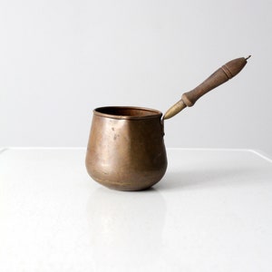 May include: A small copper pot with a wooden handle. The pot has a rounded bottom and a flared lip. The handle is attached to the pot with a brass rivet.