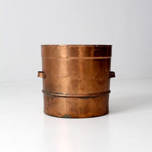 Puede incluir: Un cubo de cobre vintage con forma cilíndrica y dos asas pequeñas. El cubo tiene una pátina deslustrada, que muestra signos de edad y uso. El cobre tiene un tono marrón rojizo cálido. El diseño es simple y funcional.