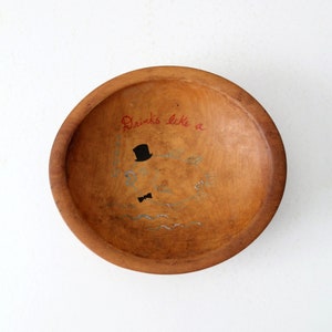 May include: A wooden bowl with a hand-painted design. The bowl is brown and has a light brown stain. The design includes a black top hat, a bow tie, and the words "Drinks like a".