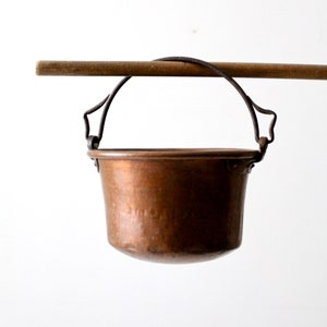 Pode incluir: Um balde de cobre com uma alça de metal pendurada em uma vara de madeira. O balde tem cerca de 15 cm de diâmetro.