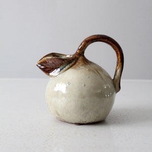 May include: A small, white ceramic pitcher with a brown handle and a brown leaf design. The pitcher has a round body and a small spout.